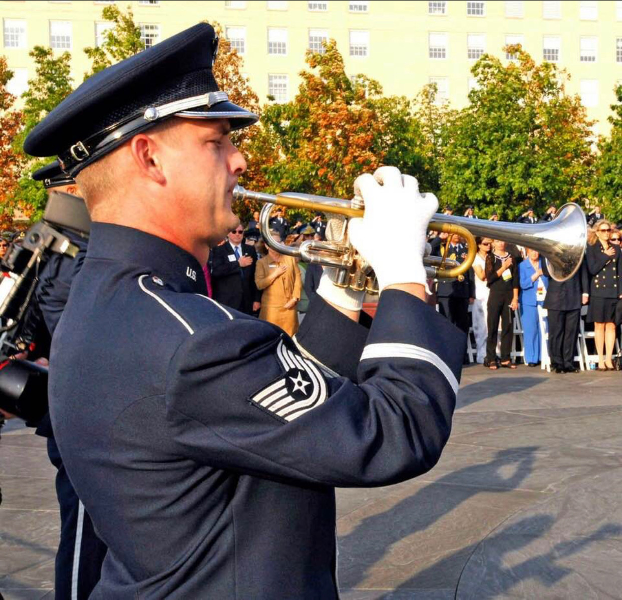 Christopher Wilson playing trumpet in Air Force Band dress uniform at an outdoor ceremony, with a uniformed band and audience behind him